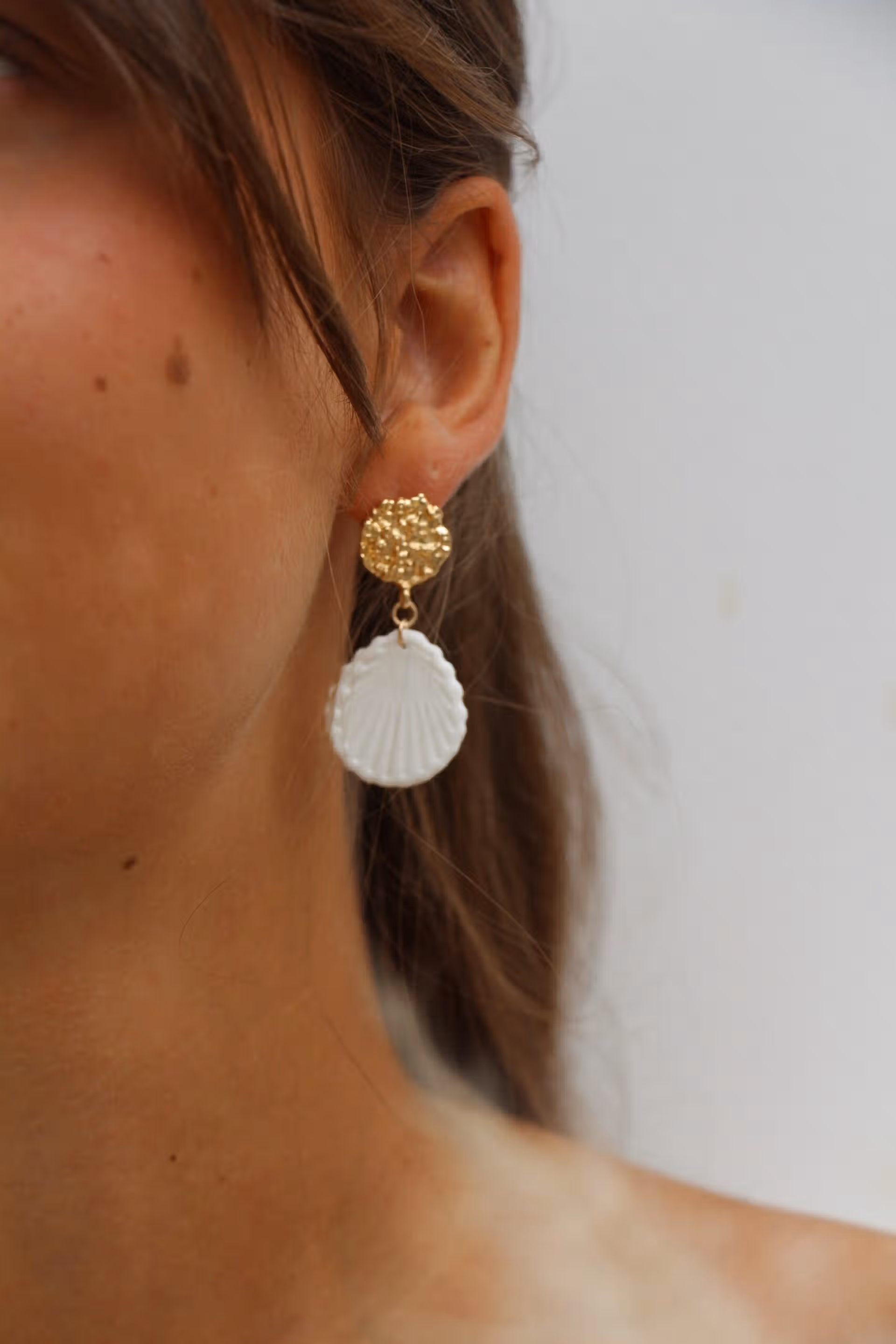 Boucles d'oreilles pendantes avec médaillon coquillage blanc et puce dorée martelée portées par une femme.